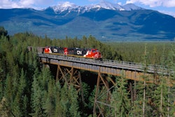 A train crosses a bridge with snow-covered mountains in the background.