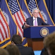 A man speaks at a podium in front of flags.
