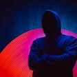 A man with no face in a dark hoodie stands in front of a dark background with neon lights.
