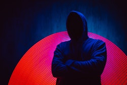 A man with no face in a dark hoodie stands in front of a dark background with neon lights.
