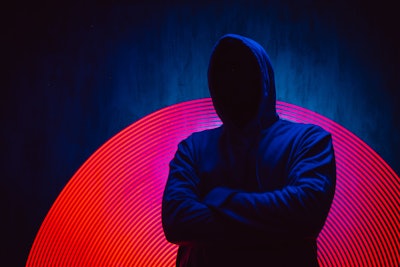 A man with no face in a dark hoodie stands in front of a dark background with neon lights.