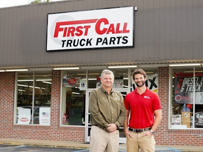 Two men stand in front of a building that says First Call Truck Parts
