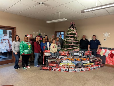 A group of people stand with a pile of toy trucks in front of a Christmas tree.