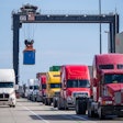 A line of trucks under a crane at a port.