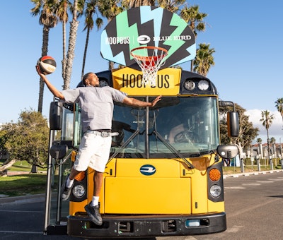 A man dunking a basketball in a hoop attached to an electric school bus from Blue Bird.