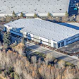 Aerial view of large warehouse complex with parking lot, surrounded by evergreen trees
