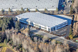 Aerial view of large warehouse complex with parking lot, surrounded by evergreen trees