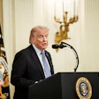 Man in dark suit speaking at presidential podium with American flag and official seal in formal government setting