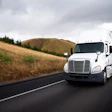 White Freightliner semi-truck driving on highway with golden hillside landscape under cloudy sky