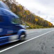 Blue truck driving on highway with autumn forest trees lining the road under cloudy sky