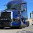 Blue Mack semi-truck with black grille parked in industrial yard under clear sky