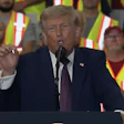Speaker at podium addressing crowd wearing safety vests with flags at political rally event