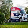 Red, white, and blue Mack semi-truck with patriotic flag design and '250 Years of Freedom' livery parked on grass with trees