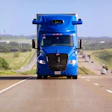 Blue semi-truck driving on a straight highway through rural farmland landscape