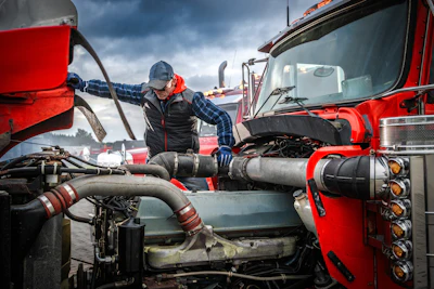 Truck driver in plaid shirt and safety gear servicing engine components on red semi-truck under cloudy sky