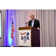 Man speaking at ATA podium with American flag and blue-lit curtains in background