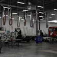 Industrial truck maintenance facility with white and red commercial vehicles suspended on lift systems under bright overhead lighting.