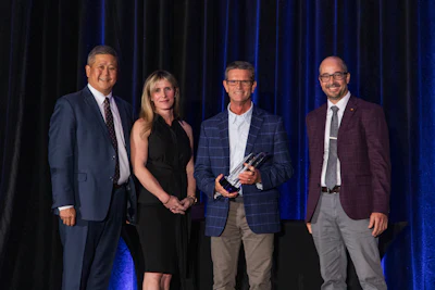 Four professionals stand together at formal event, man in center holding award against blue curtain backdrop
