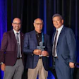 Three men in business attire holding an award at a formal event with blue stage lighting