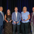 Five professionals holding awards at a formal event with blue curtain backdrop