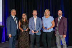 Five professionals holding awards at a formal event with blue curtain backdrop