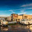 Commercial warehouse facility with fleet of white service vehicles parked outside on wet pavement under blue sky