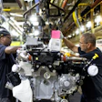 Two automotive workers assembling an engine component on a factory assembly line using overhead lifting equipment.