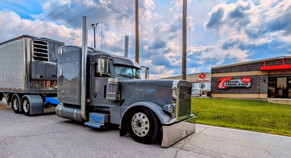 Black and blue Peterbilt semi-truck with refrigerated trailer parked at industrial facility under cloudy sky