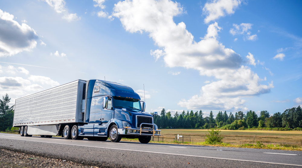 Blue and white semi-truck with refrigerated trailer driving on highway through rural landscape with forest and clear sky