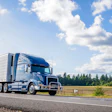 Blue and white semi-truck with refrigerated trailer driving on highway through rural landscape with forest and clear sky