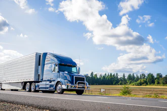 Blue and white semi-truck with refrigerated trailer driving on highway through rural landscape with forest and clear sky