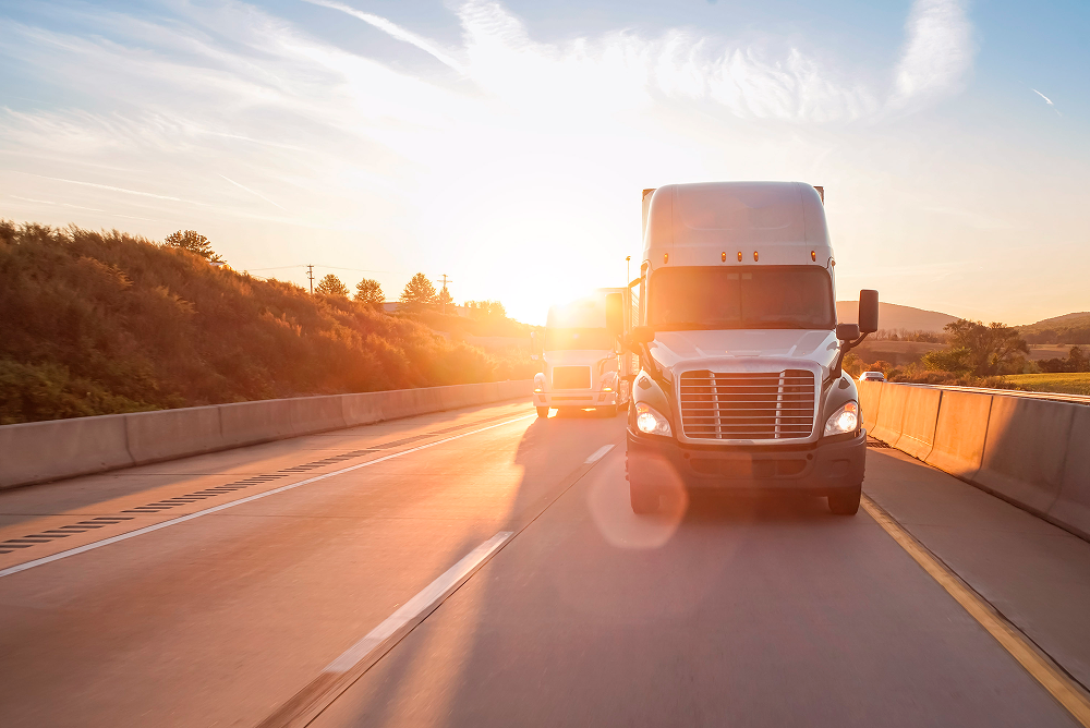 Semi-trucks driving on highway at golden sunset with hills and guardrails visible