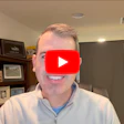 Man in office with YouTube play button overlay on face, sitting at desk with wooden shelving and framed photos
