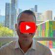 Man smiling at camera with YouTube play button overlay, city skyline and colorful bookshelf visible through office window
