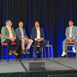 Four men seated on stage during a panel discussion with blue curtain backdrop at a conference event