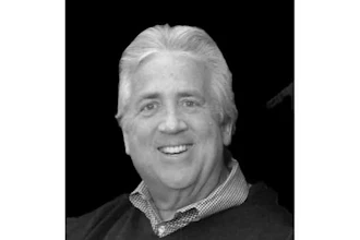 Professional headshot of a smiling middle-aged man with white hair wearing a dark sweater and checkered shirt collar