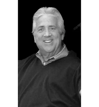 Professional headshot of a smiling middle-aged man with white hair wearing a dark sweater and checkered shirt collar