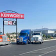 Kenworth truck dealership with three semi-trucks parked under red and white signage against mountain backdrop