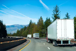 Semi-trucks driving on a curved highway through pine forest with mountains visible in the distance under clear blue sky