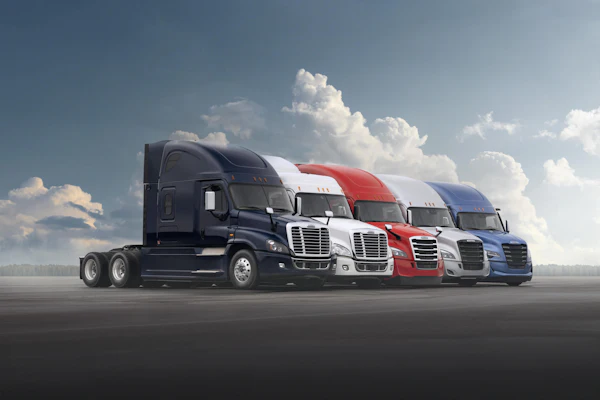 Row of five commercial semi-trucks in navy, white, red, and blue colors parked on asphalt against a cloudy sky