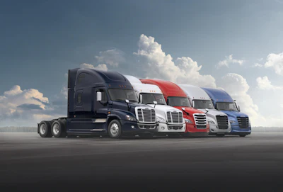 Row of five commercial semi-trucks in navy, white, red, and blue colors parked on asphalt against a cloudy sky