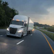 White semi-truck with illuminated headlights driving on a multi-lane highway during dusk with green trees and fencing alongside the road.