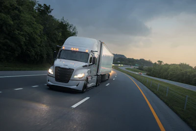 White semi-truck with illuminated headlights driving on a multi-lane highway during dusk with green trees and fencing alongside the road.