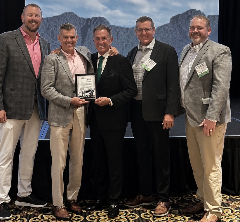 Five men in business attire pose together at an event while holding an award plaque, with mountains visible in the background.