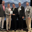 Five men in business attire pose together at an event while holding an award plaque, with mountains visible in the background.