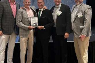 Five men in business attire pose together at an event while holding an award plaque, with mountains visible in the background.