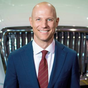 Professional headshot of a smiling man in navy blazer and burgundy tie against a blurred background