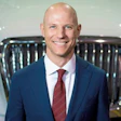 Professional headshot of a smiling man in navy blazer and burgundy tie against a blurred background