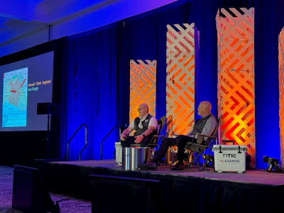 Two speakers engage in panel discussion on stage with blue lighting and geometric orange backdrop panels at a professional conference event.