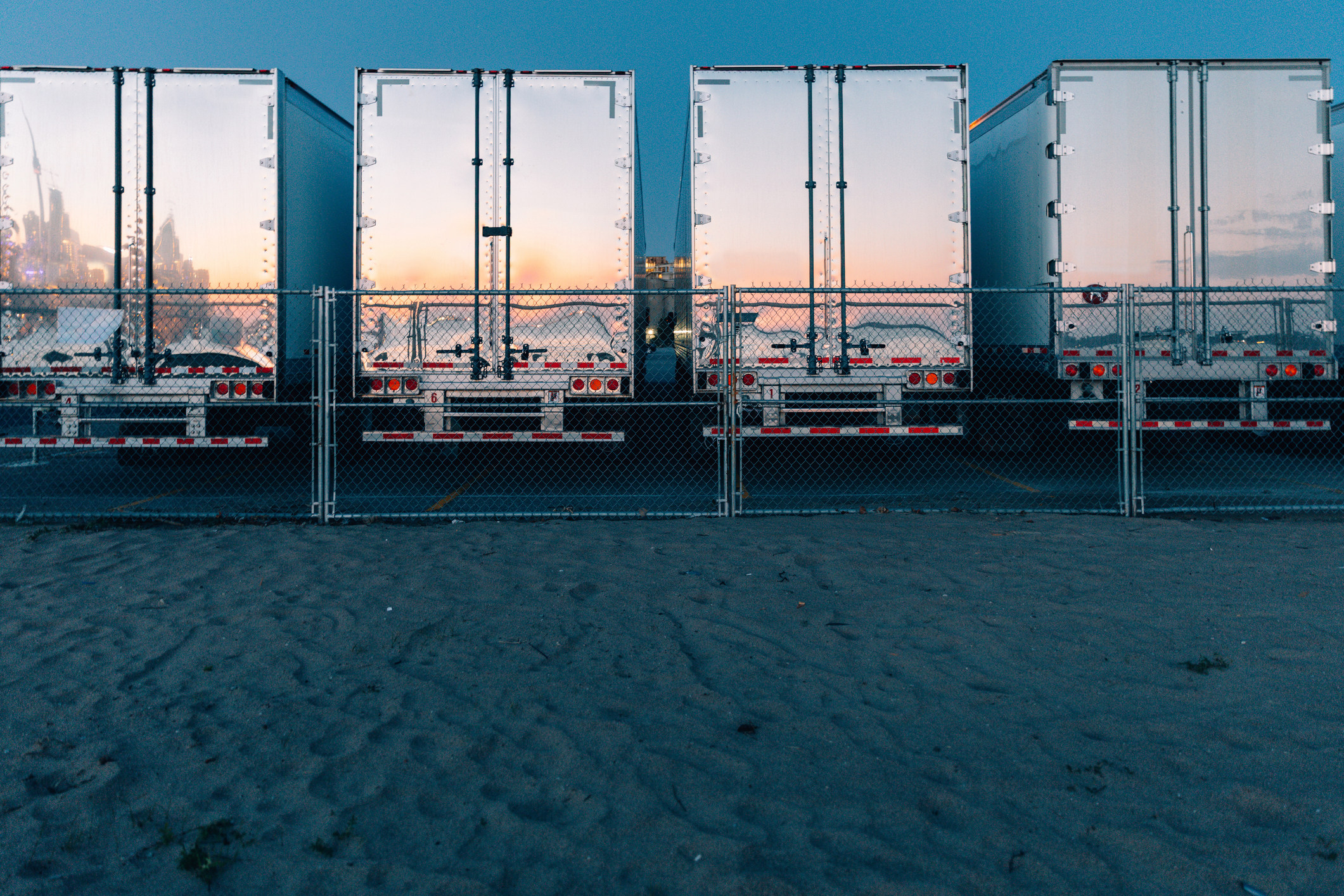 Row of industrial shipping containers at a waterfront dock with city skyline visible through open doors at sunset